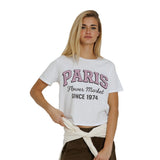 A blonde woman modeling a white cropped graphic T-shirt with 'PARIS Flower Market SINCE 1974' printed in pink and black, paired with dark brown pants and a cream tie-waist sash, against a plain white background.