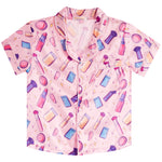 A short-sleeve button-up pajama shirt in light pink with an all-over print of colorful makeup items, including lipsticks, brushes, compacts, and palettes, shown on a plain white background.