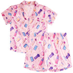 A two-piece girls' pajama set featuring a short-sleeve button-up top and matching shorts in light pink, both printed with a colorful cosmetic-themed pattern of lipsticks, brushes, powders, and makeup containers.