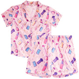 A two-piece girls' pajama set featuring a short-sleeve button-up top and matching shorts in light pink, both printed with a colorful cosmetic-themed pattern of lipsticks, brushes, powders, and makeup containers.