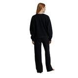 A woman with long light brown hair is shown from the back wearing a matching black sweatshirt and wide-leg sweatpants set, standing against a solid white background.