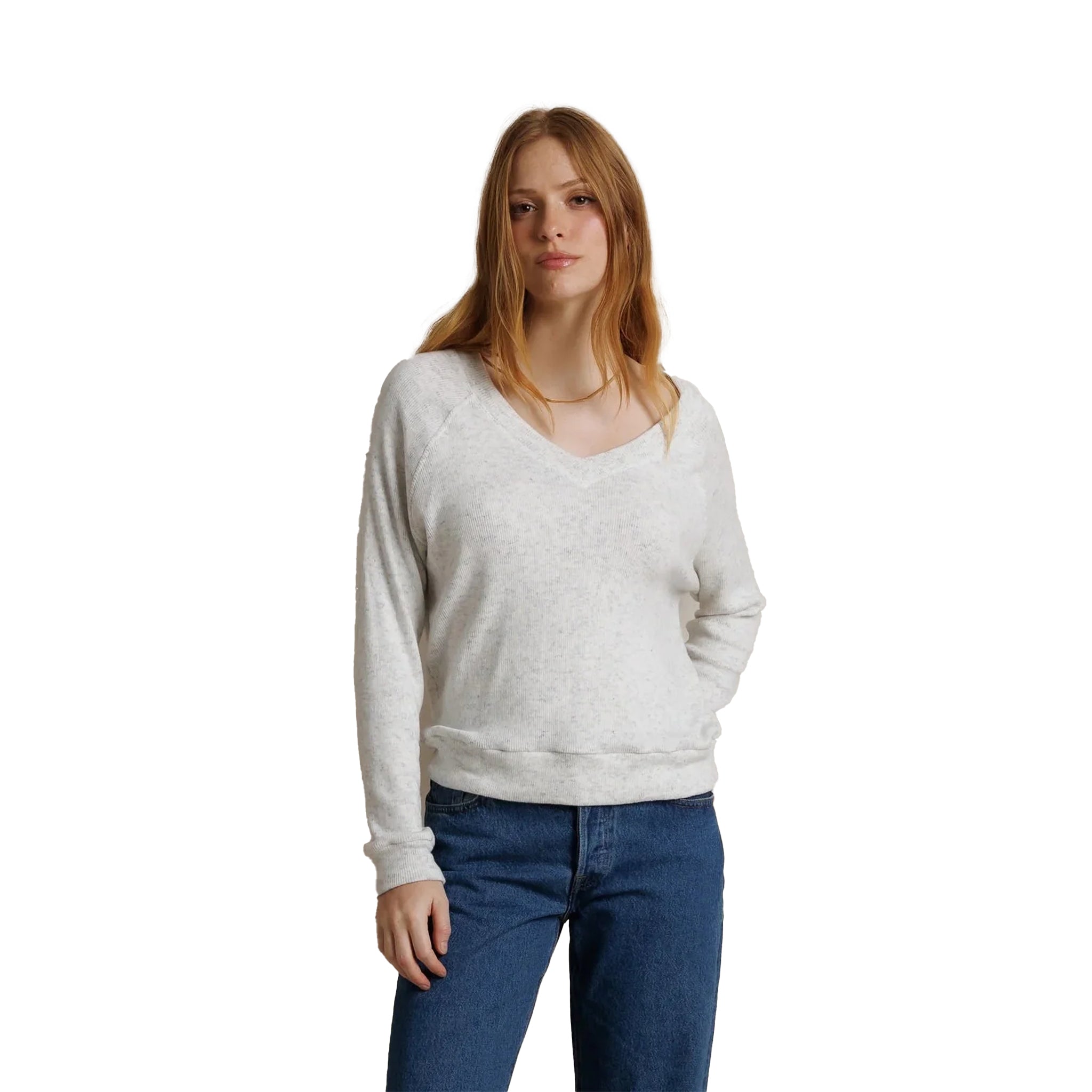 A woman with red hair wearing a light heather gray v-neck sweater and blue jeans, standing against a solid white background.