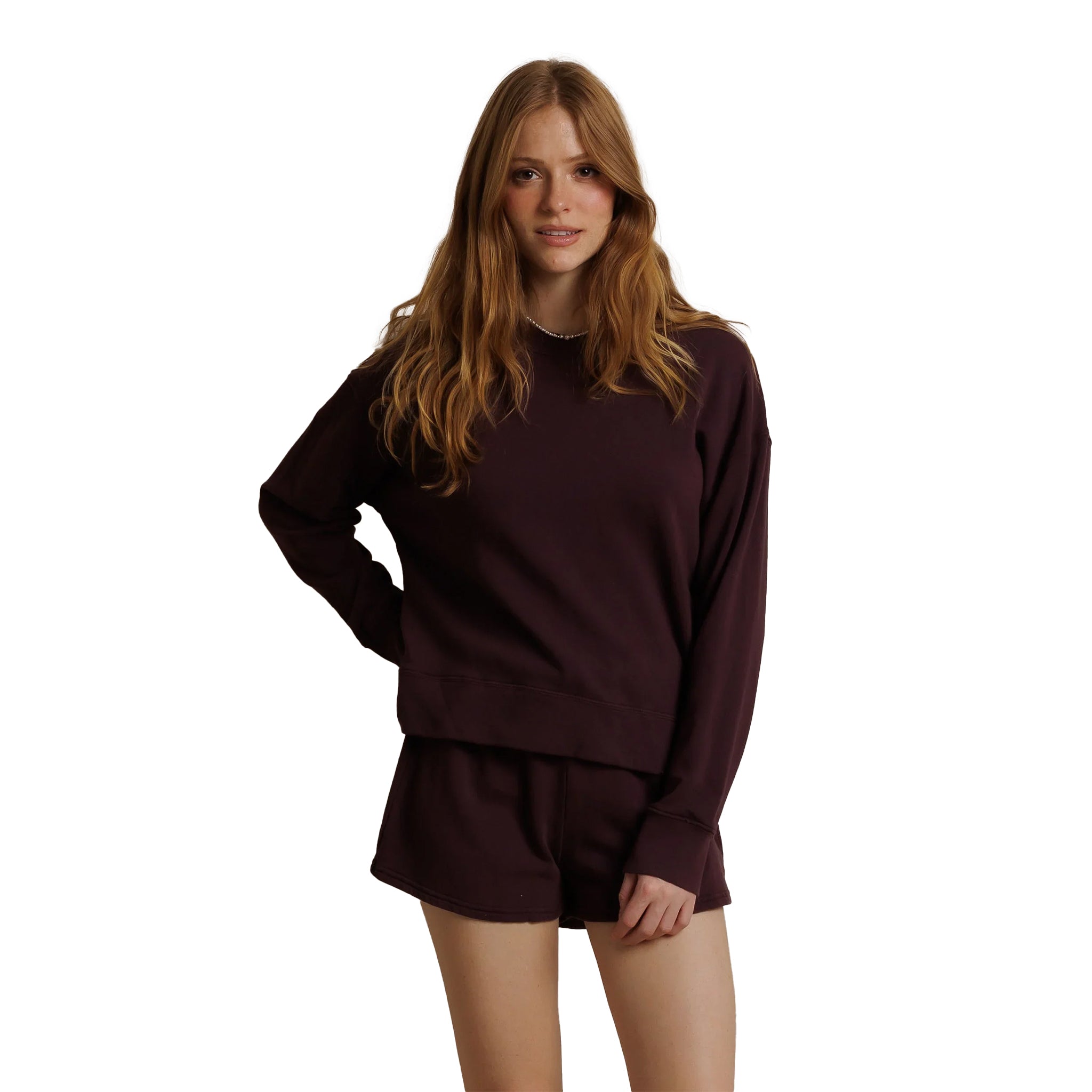 A woman modeling a matching dark plum crewneck sweatshirt and shorts lounge set against a solid white background.