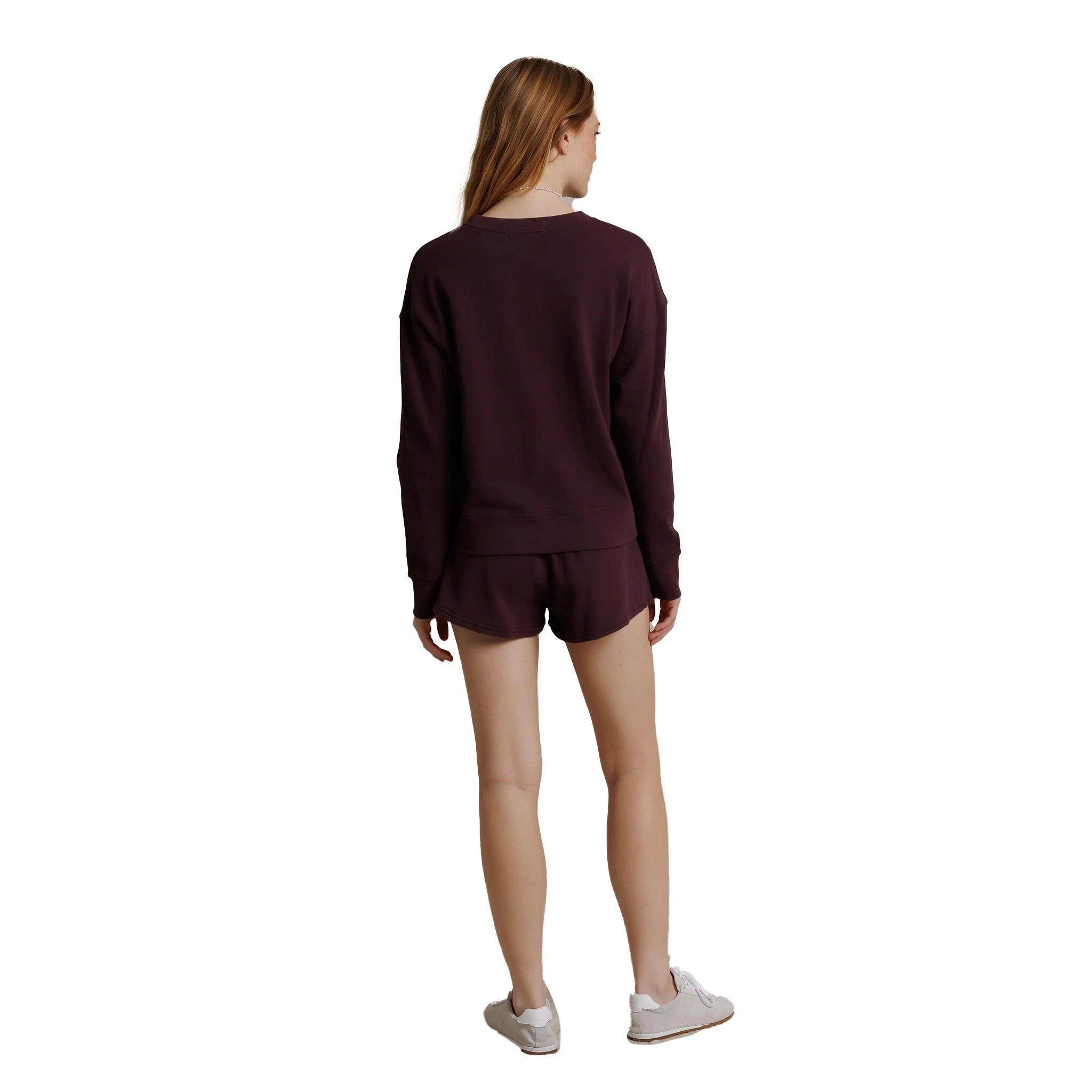A woman viewed from behind wearing a matching dark burgundy long-sleeve top and shorts set, paired with light gray sneakers, against a plain white background.