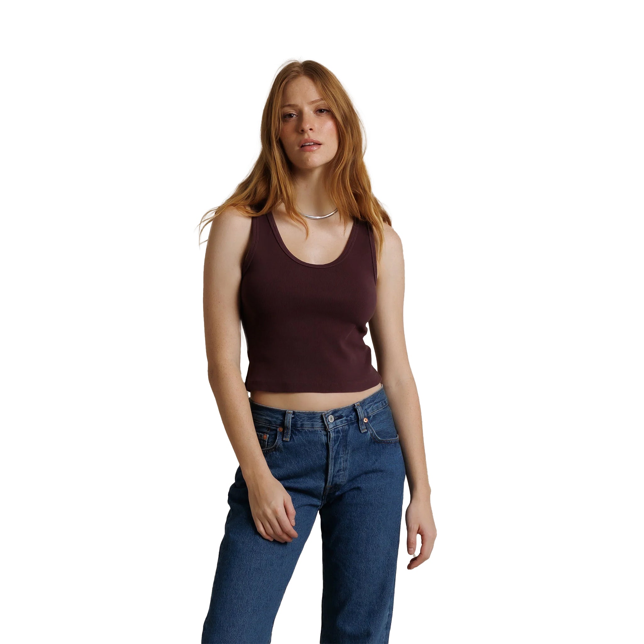 A woman with red hair wearing a dark maroon ribbed scoop-neck tank top and blue jeans, standing against a solid white background.