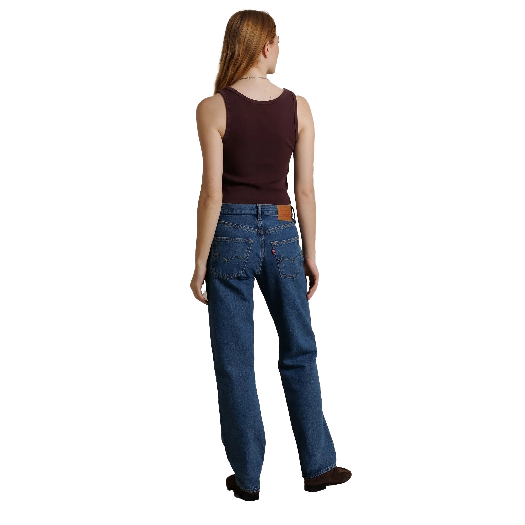 A woman shown from the back wearing blue jeans, a dark brown sleeveless top, and brown shoes against a plain white background.