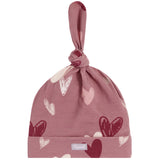 A dusty rose knit baby hat with a knotted top and a folded cuff, decorated with a pattern of hearts in various shades of pink, red, and white. A small silver brand tag is visible on the cuff.