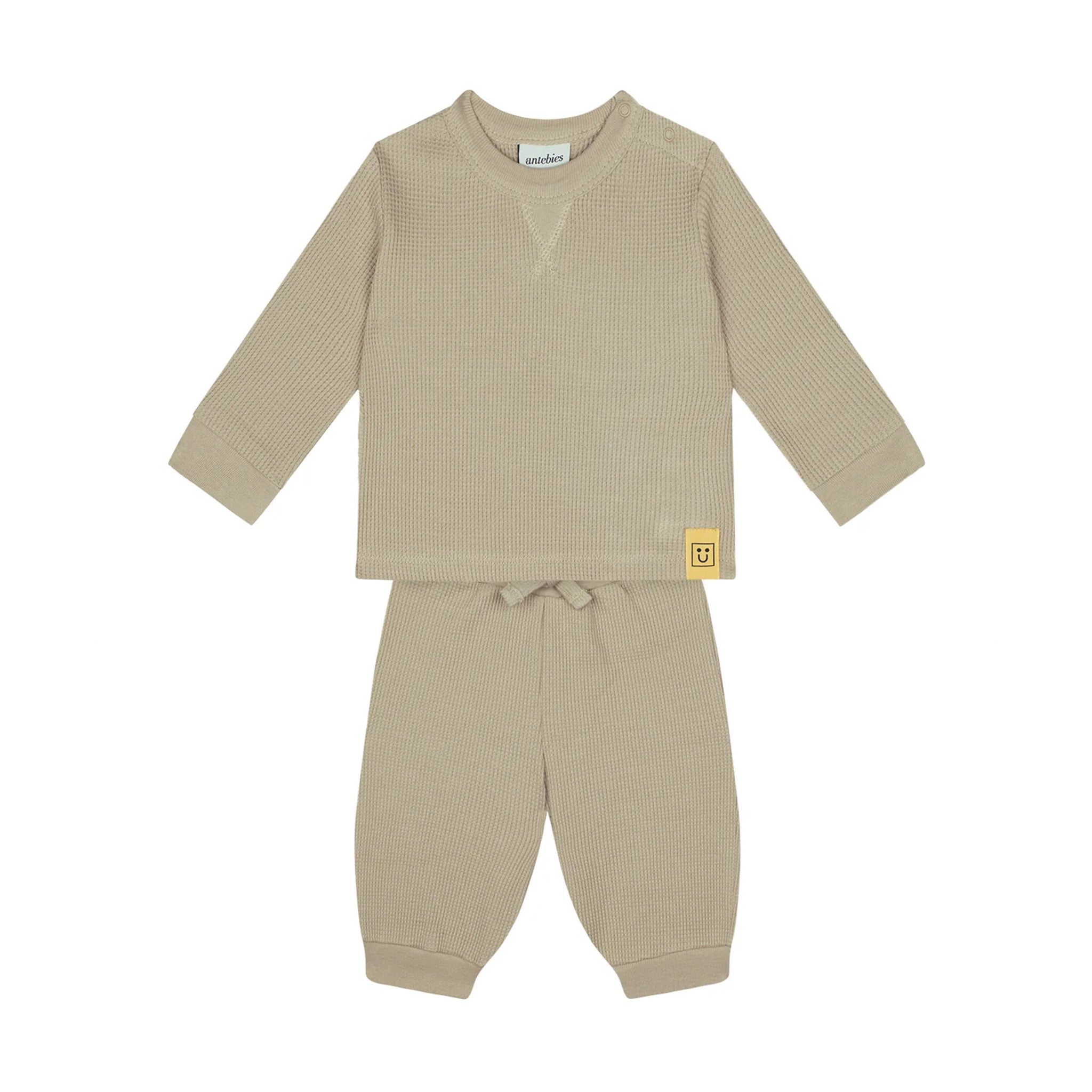 A two-piece baby loungewear set in a warm beige waffle-knit fabric, featuring a long-sleeve top with a small yellow brand patch on the sleeve and matching pants with a drawstring waist.