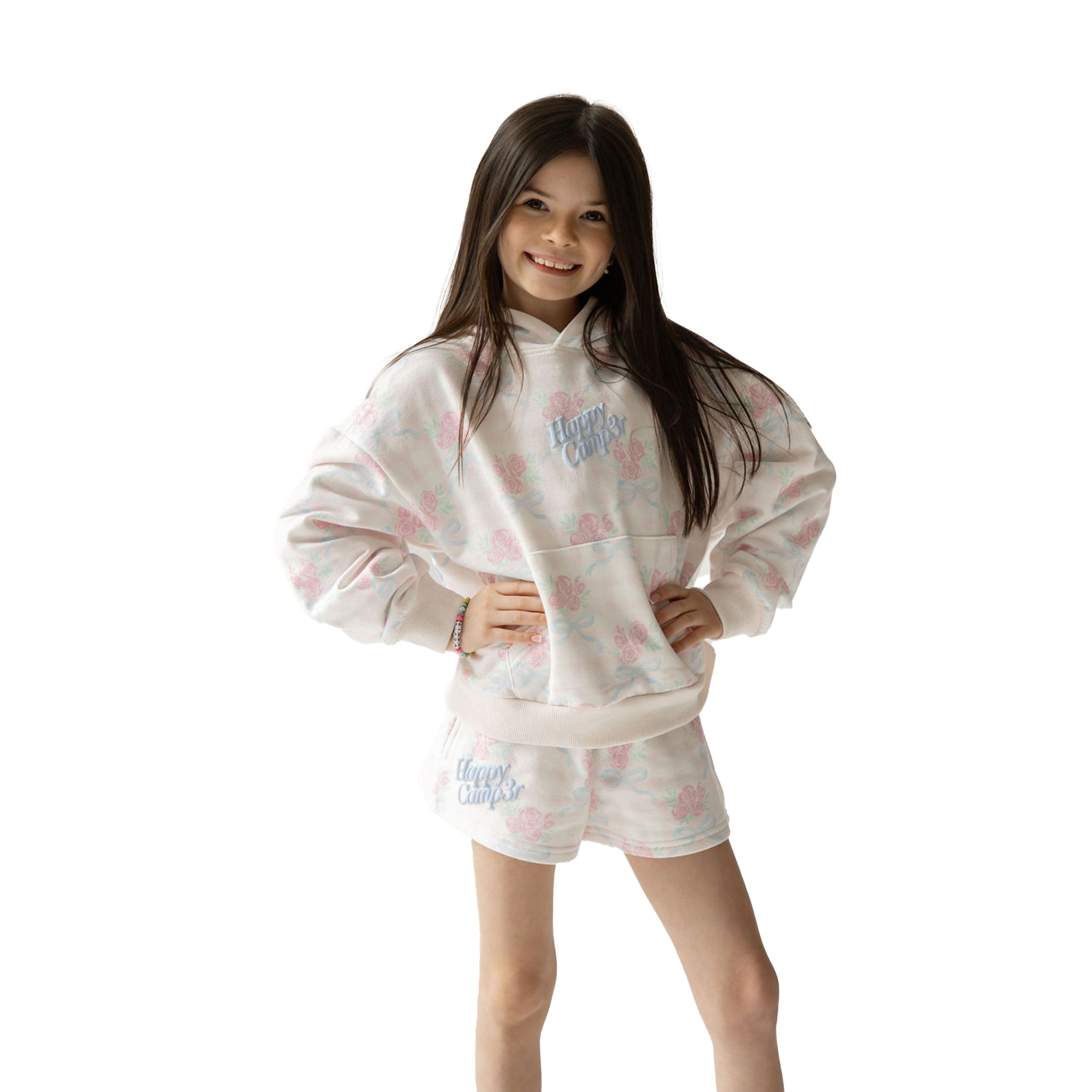 Young girl wearing a white hoodie and shorts set with pink floral patterns and blue 'happy camper' text on a white background