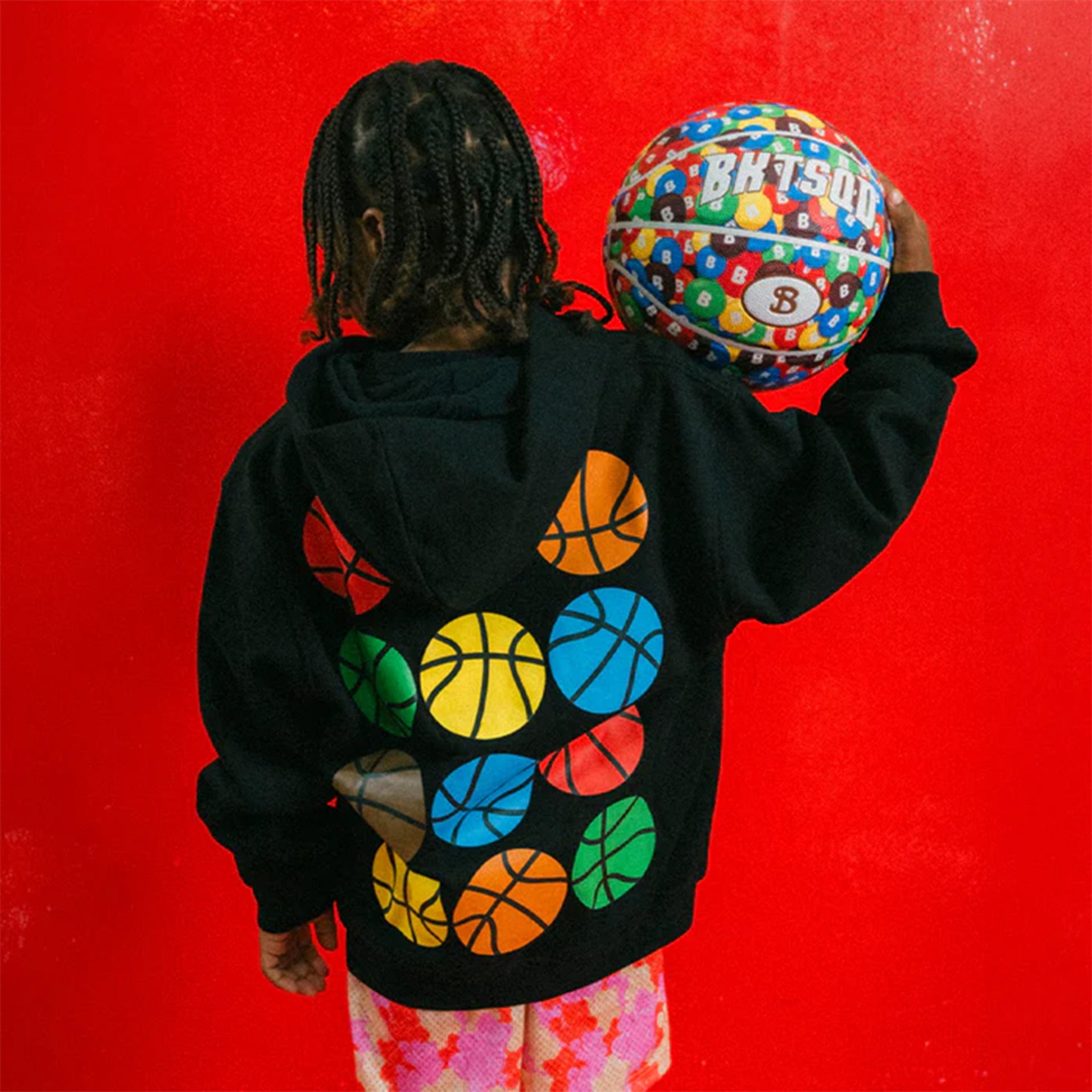 Child wearing a black hoodie with colorful basketballs on the back and gummy bear patterned shorts, holding a multicolored basketball against a red background.