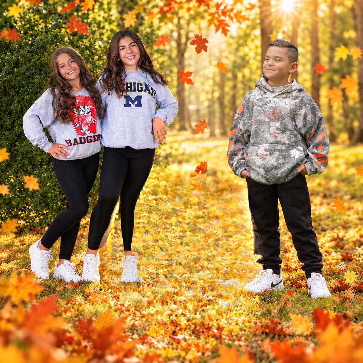 Three children standing outdoors in a fall park with fallen leaves and autumn trees in the background. The girl on the left wears a gray sweatshirt with a red and white Badgers graphic, the middle child wears a light gray sweatshirt with a blue Michigan 'M' logo, and the boy on the right wears a gray camouflage hoodie with orange accents.