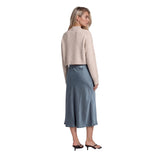 A woman shown from the back wearing a light beige cropped cardigan and a slate blue midi skirt, paired with black mule sandals, against a plain white background.