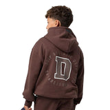 A child model shown from the back wearing a dark brown cropped hoodie with a large white outlined letter 'D' and the words 'DRIP ATHLETIC DEPT. ESTABLISHED 2023' printed on the back, paired with matching brown sweatpants against a plain white background.
