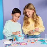 A boy and a girl are shown participating in a colorful craft or painting activity at a table. The girl holds a small painted object, while the boy mixes paint in a light blue bowl.